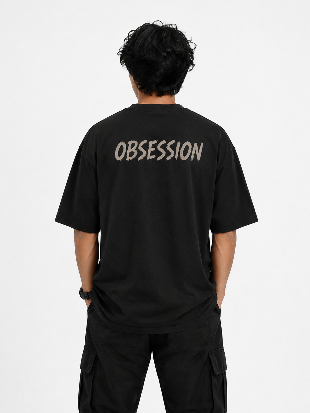 A man viewed from behind wearing a black t-shirt with the word 'OBSESSION' printed in a light beige stencil font across the upper back, paired with black cargo pants against a plain white studio background.