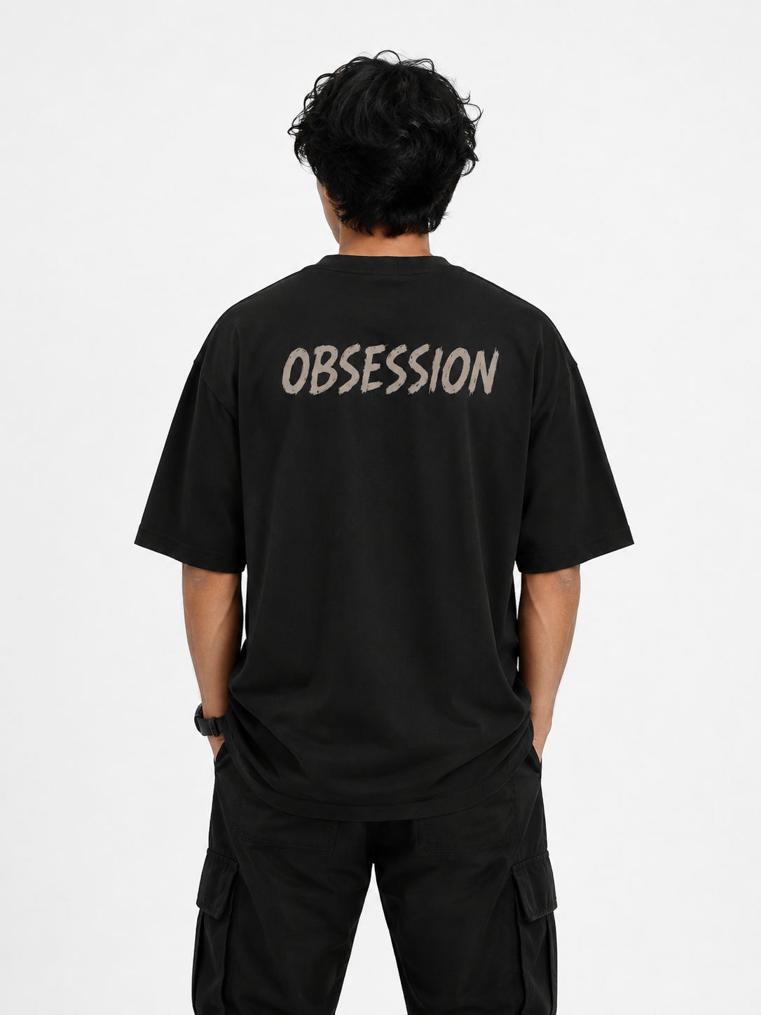 A man viewed from behind wearing a black t-shirt with the word 'OBSESSION' printed in a light beige stencil font across the upper back, paired with black cargo pants against a plain white studio background.